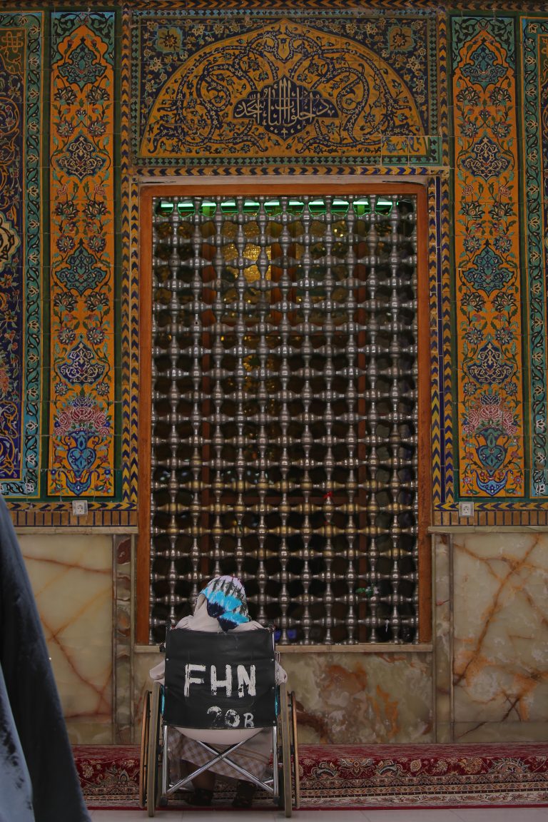 یک زائر روی ویلچر در مقابل پنجره مشبک ایوان ناودان طلا