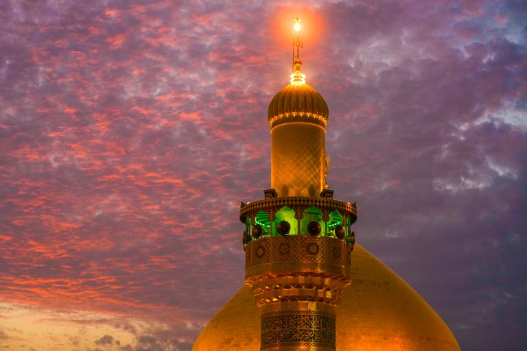 گلدسته حرم امام علی در مقابل گنبد و ابرهای نارنجی و کبود طلوع