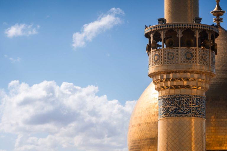 نمای نزدیک مناره حرم مطهر امیرالمؤمنین علی و میناکاری‌های آن با آسمان آبی و ابرهای سفید