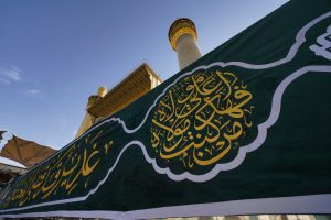 نمای نزدیک کتیبه سبز غدیری با گلدسته حرم علوی در پس‌زمینه