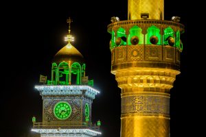 نمای نزدیک گلدسته و مناره ساعت حرم علوی با نورهای سبز و طلایی