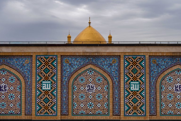 نمایی از کاشی‌کاری‌های هندسی حرم علوی با گنبد طلایی در پس‌زمینه