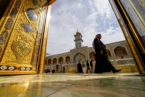 زائری در صحن حرم مطهر امیرالمؤمنین با درگاه طلایی و گلدسته در پس‌زمینه